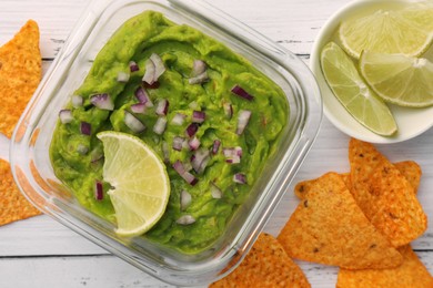 Bowl of delicious guacamole with onion, lime and nachos chips on white wooden table, flat lay