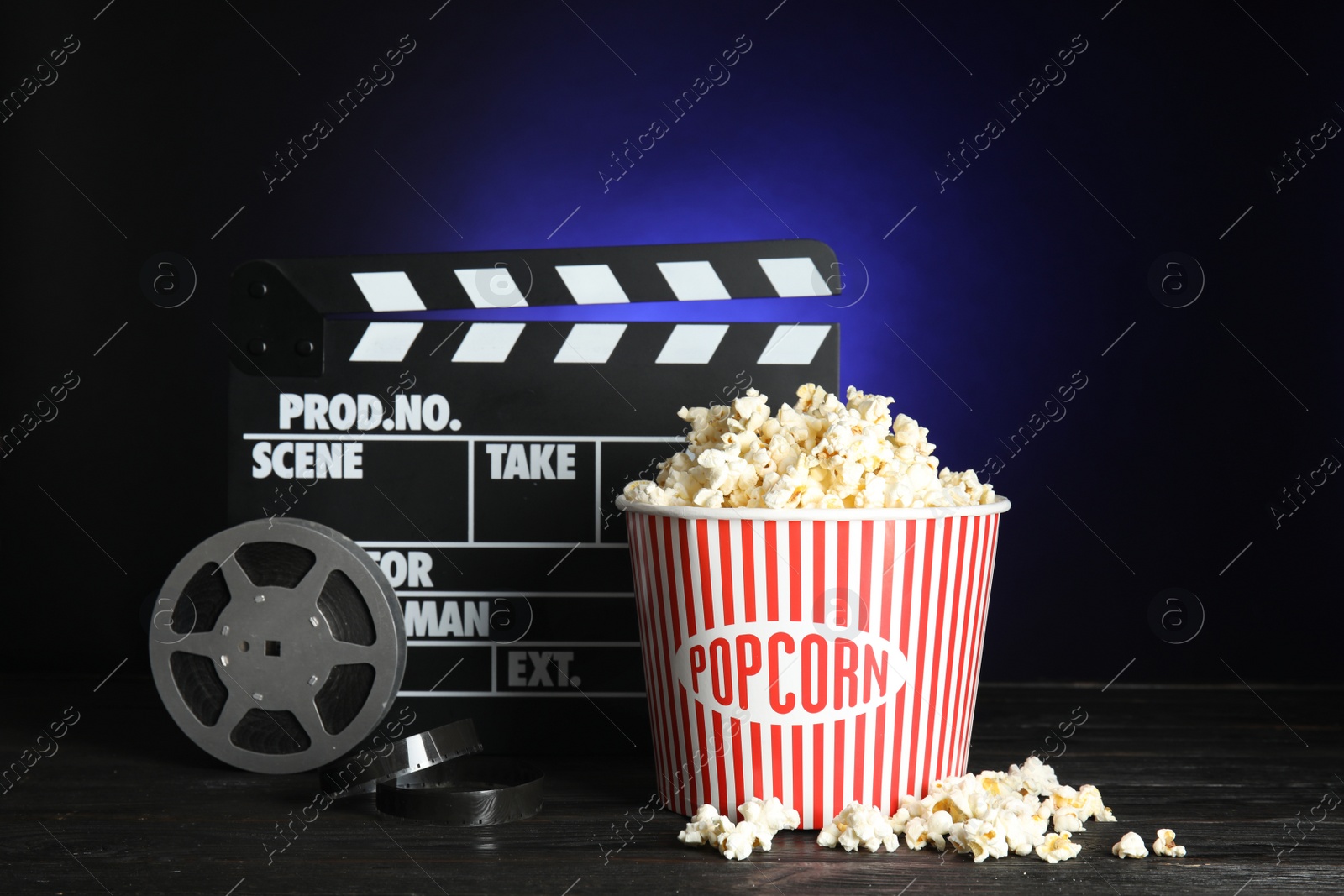 Photo of Clapperboard, reel and popcorn bucket on table against dark background. Watching cinema