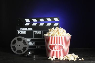 Clapperboard, reel and popcorn bucket on table against dark background. Watching cinema