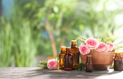 Bottles with essential oils, roses and rosemary on wooden table against blurred green background. Space for text