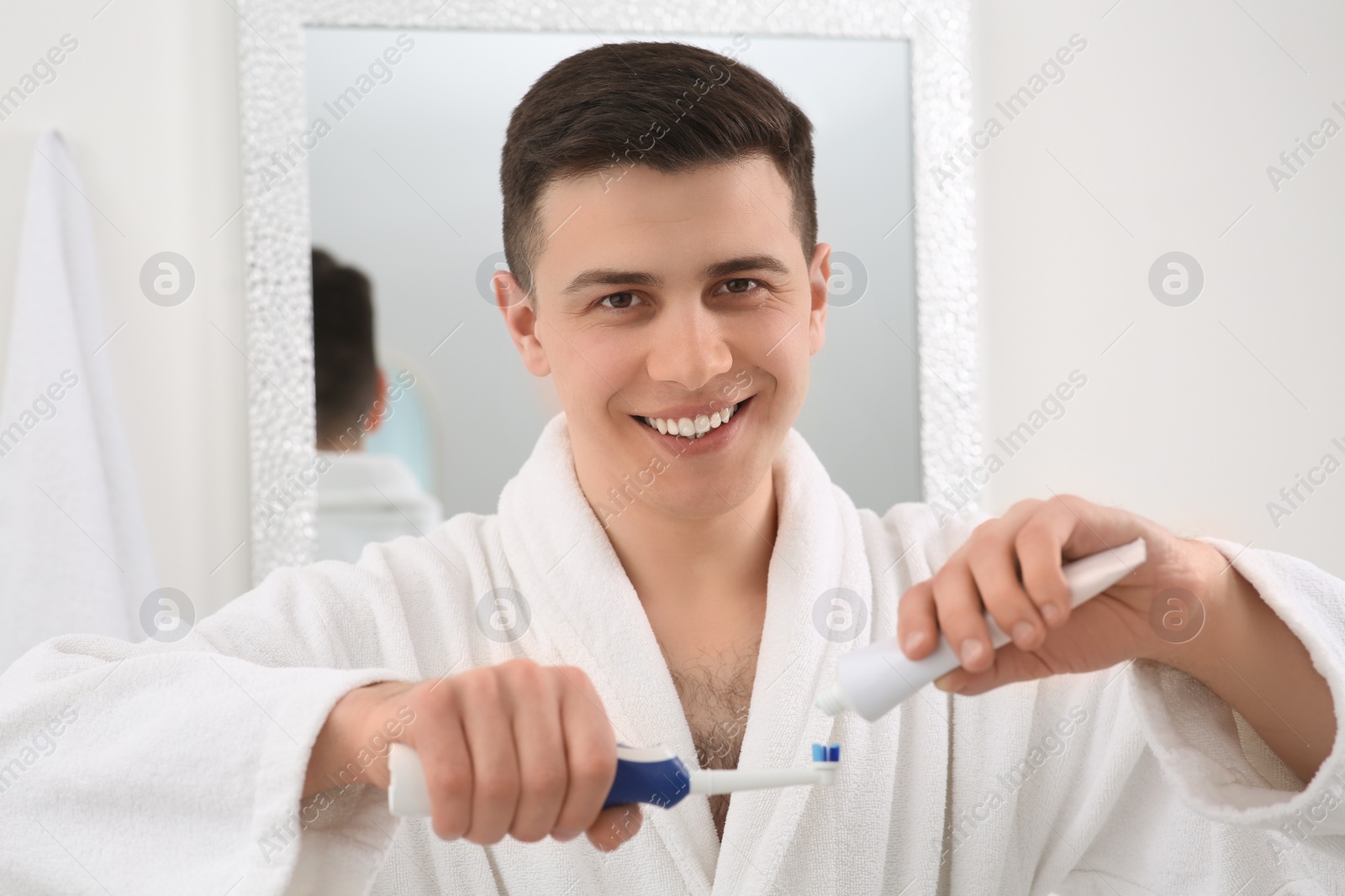 Photo of Happy man squeezing toothpaste from tube onto electric toothbrush in bathroom