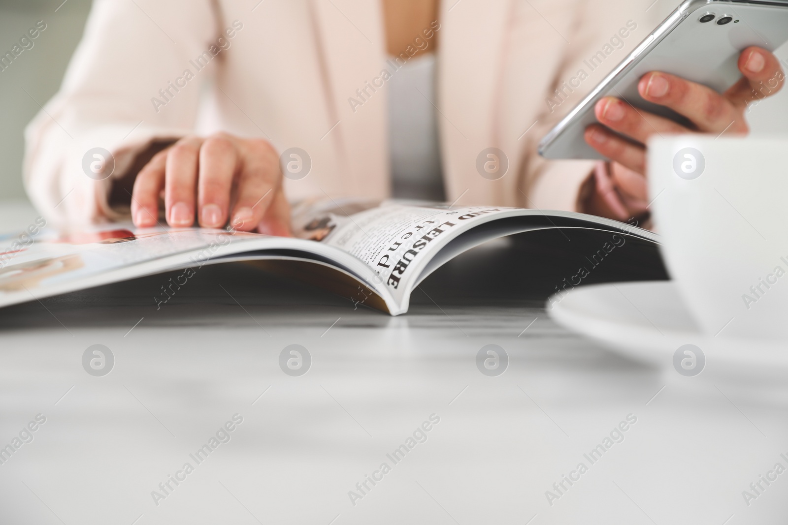 Photo of Woman with mobile phone reading magazine at white table, closeup