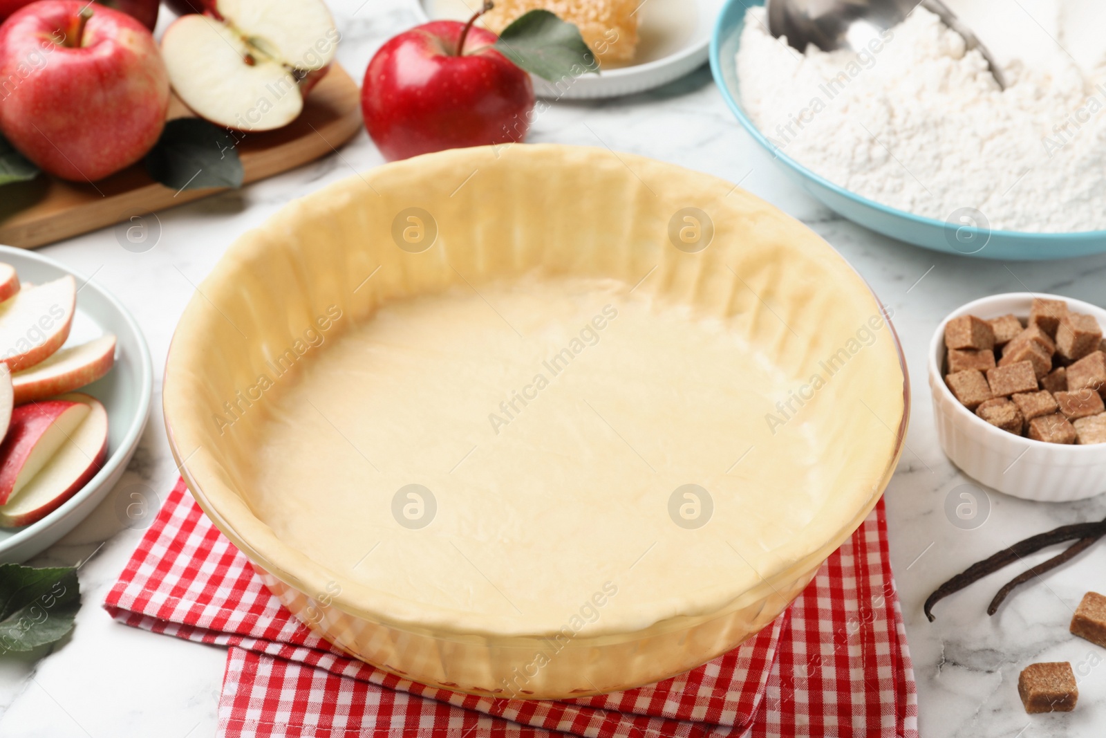 Photo of Baking dish with raw dough for apple pie and ingredients on white marble table