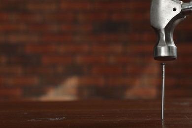 Photo of Hammering metal nail on wooden table against brick wall, closeup. Space for text