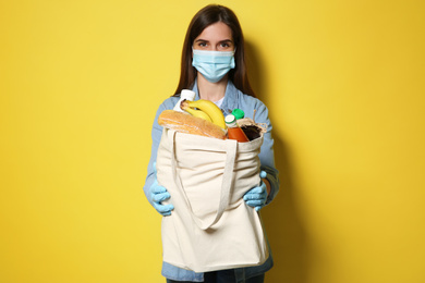 Photo of Female volunteer in protective mask and gloves with products on yellow background. Aid during coronavirus quarantine