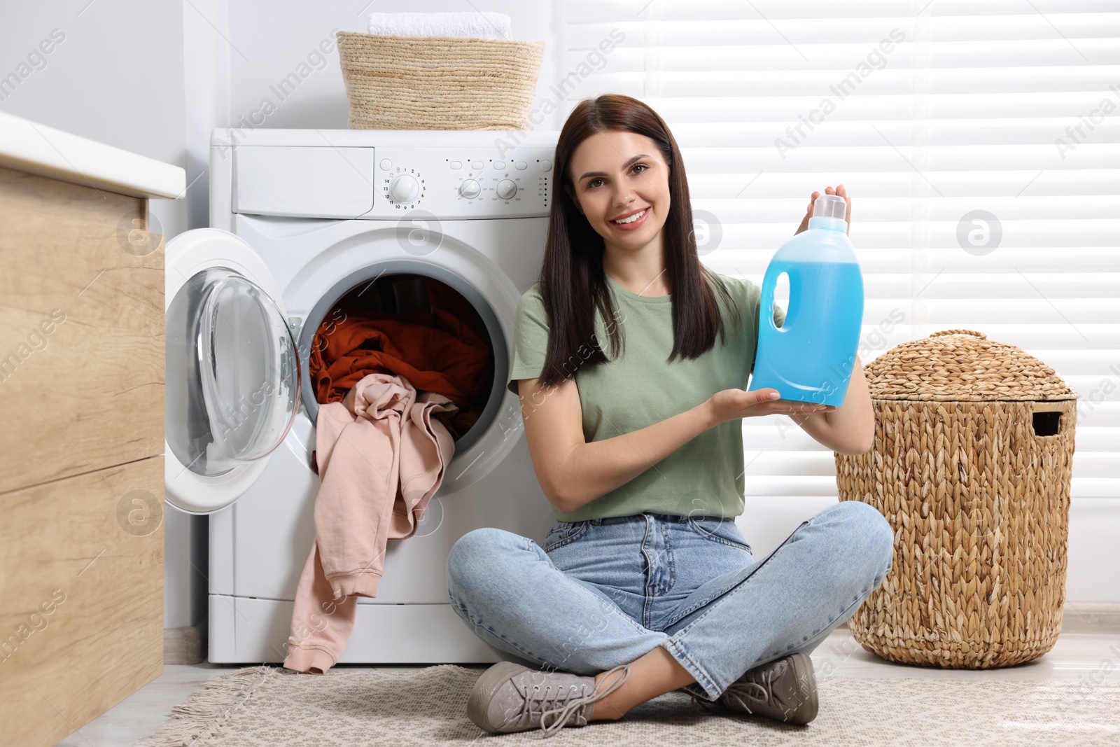 Photo of Woman sitting on floor near washing machine and holding fabric softener in bathroom