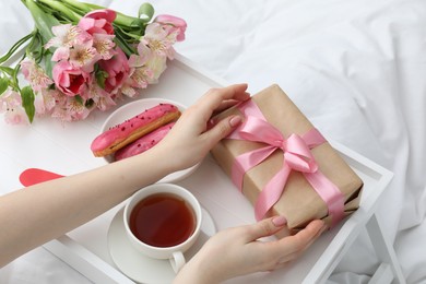 Photo of Tasty breakfast served in bed. Woman with gift box, tea, eclairs and flowers at home, above view