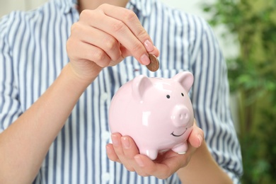 Photo of Woman putting coin into piggy bank indoors, closeup