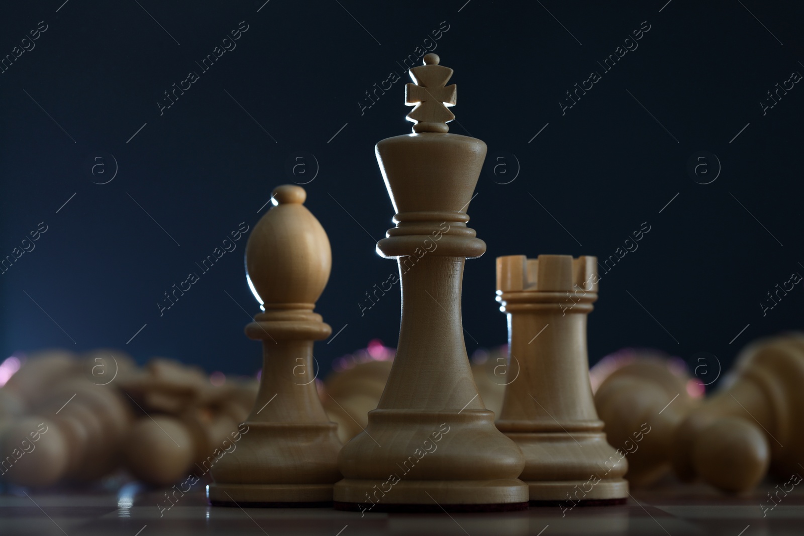 Photo of Wooden king, rook and bishop in front of fallen chess pieces on checkerboard against dark background, closeup