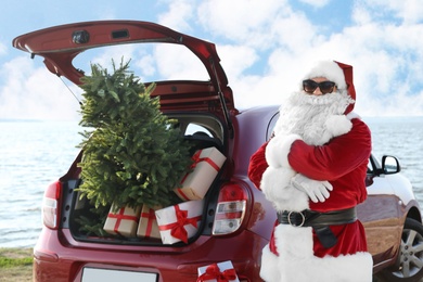 Photo of Authentic Santa Claus near red car with gift boxes and Christmas tree on beach