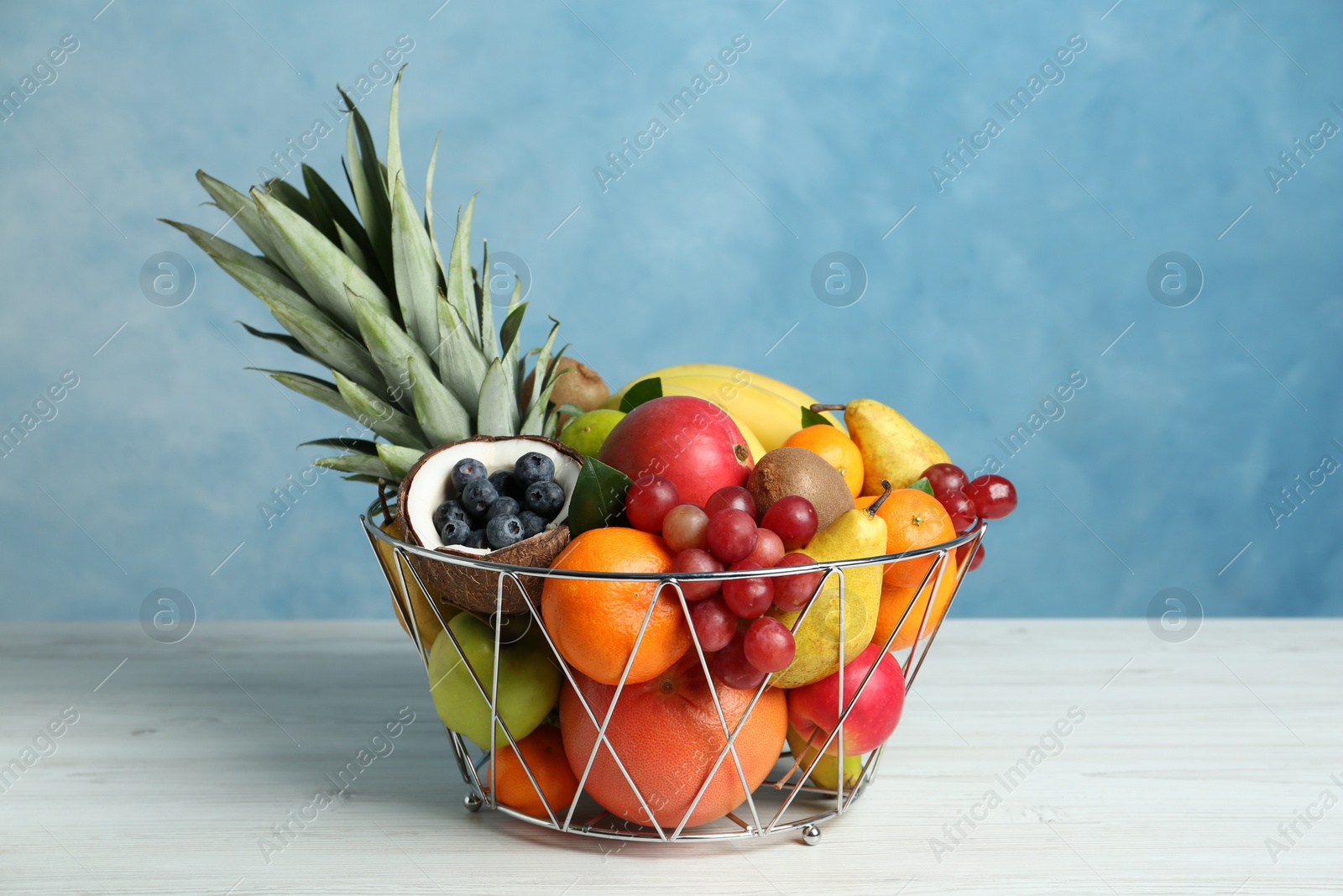 Photo of Assortment of fresh exotic fruits in metal basket on white wooden table against light blue background