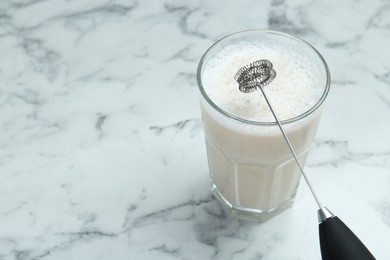 Photo of Mini mixer (milk frother) and tasty cappuccino in glass on white marble table, closeup. Space for text