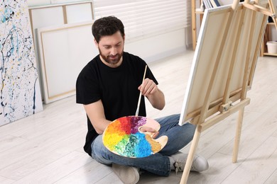Photo of Man with brush and palette painting in studio. Using easel to hold canvas