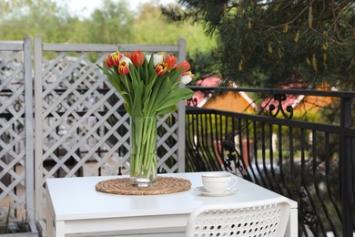 Beautiful colorful tulips in glass vase and cup of drink on white table at balcony