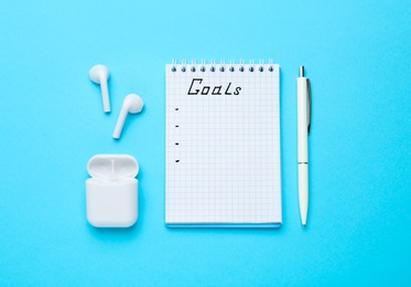 Photo of Notebook with goal list, earphones and pen on blue background, flat lay