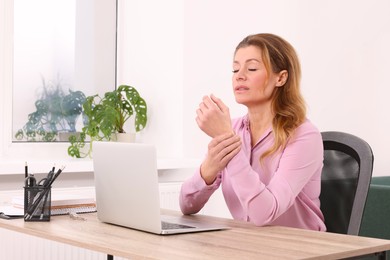 Photo of Woman suffering from pain in wrist while using laptop at wooden table indoors. Arthritis symptoms