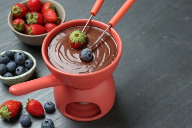 Photo of Dipping fresh berries into fondue pot with melted chocolate at grey table, closeup. Space for text