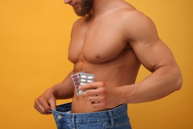 Photo of Athletic man with pills wearing big jeans on orange background, closeup. Weight loss