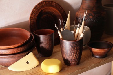 Photo of Set of different crafting tools and clay dishes on wooden rack in workshop