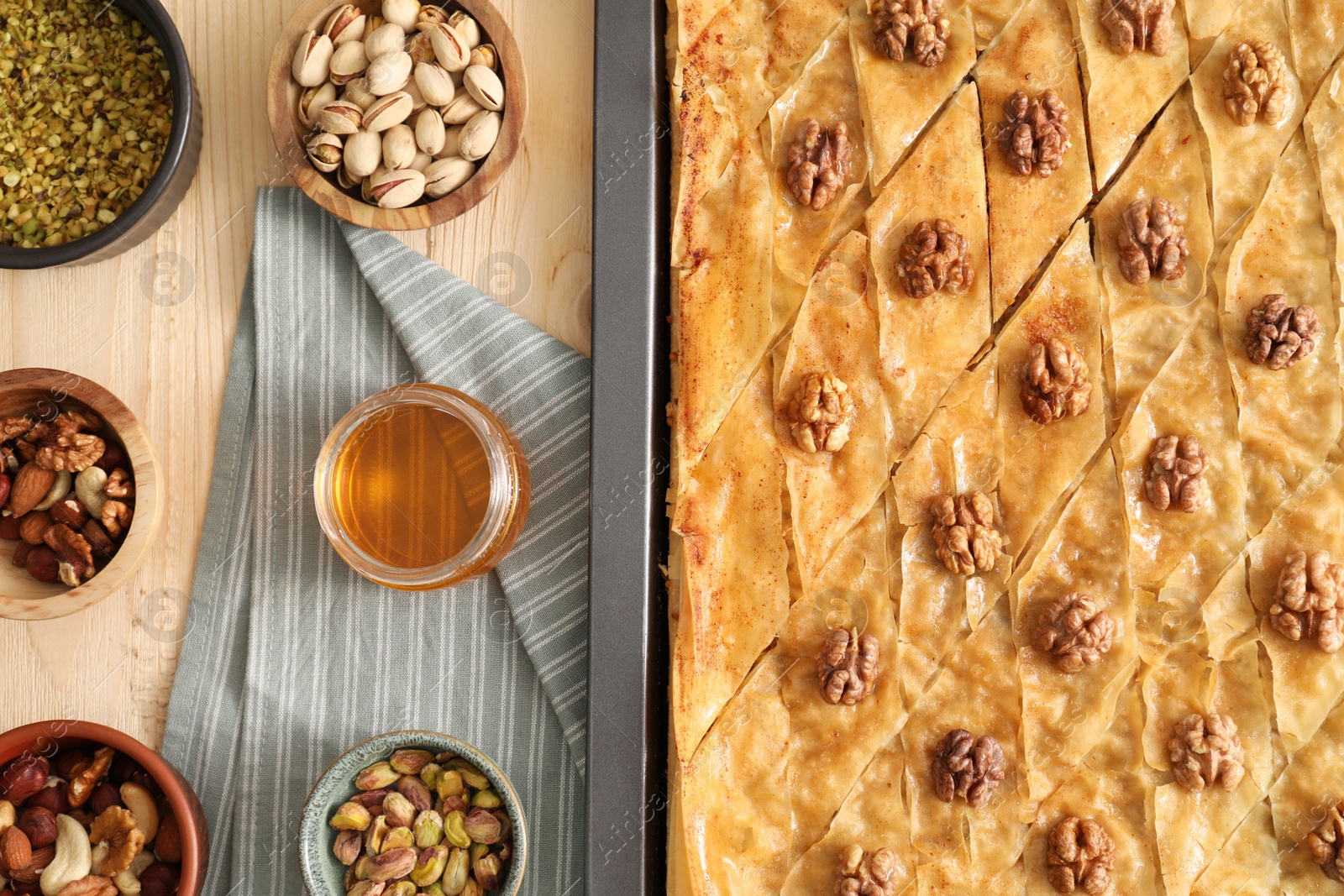 Photo of Delicious baklava with walnuts in baking pan, honey and nuts on wooden table, flat lay