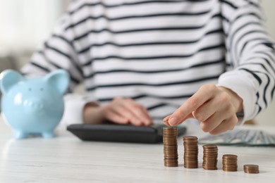 Photo of Financial savings. Woman stacking coins while using calculator at white wooden table, closeup