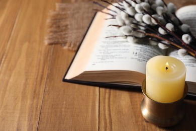 Photo of Burning candle, Bible and willow branches on wooden table, space for text