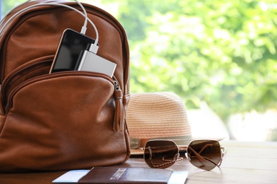 Photo of Sunglasses, passport, hat and backpack with gadgets on wooden table, space for text. Charging electronics during travel