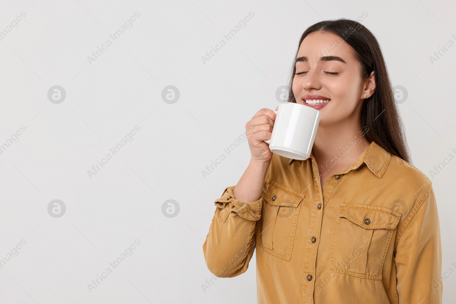Photo of Happy young woman drinking beverage from white ceramic mug on light grey background, space for text
