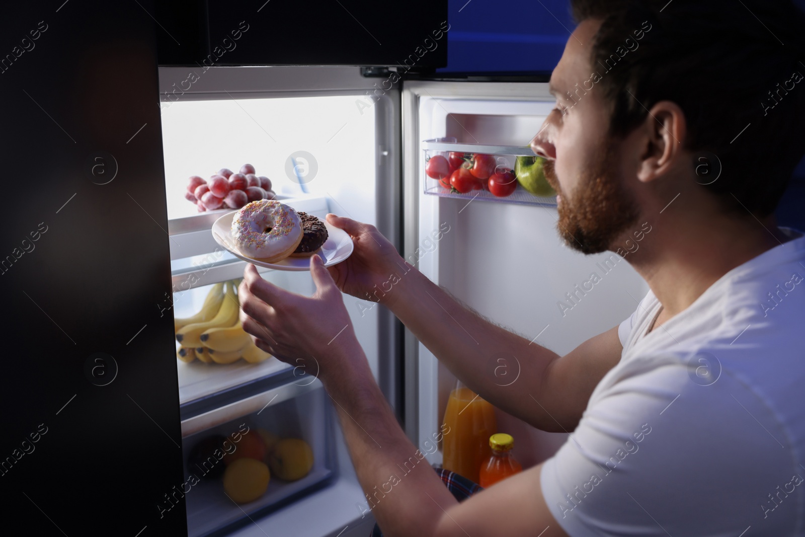 Photo of Man taking plate with donuts from refrigerator in kitchen at night. Bad habit