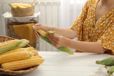 Photo of Woman husking corn cob at white wooden table, closeup