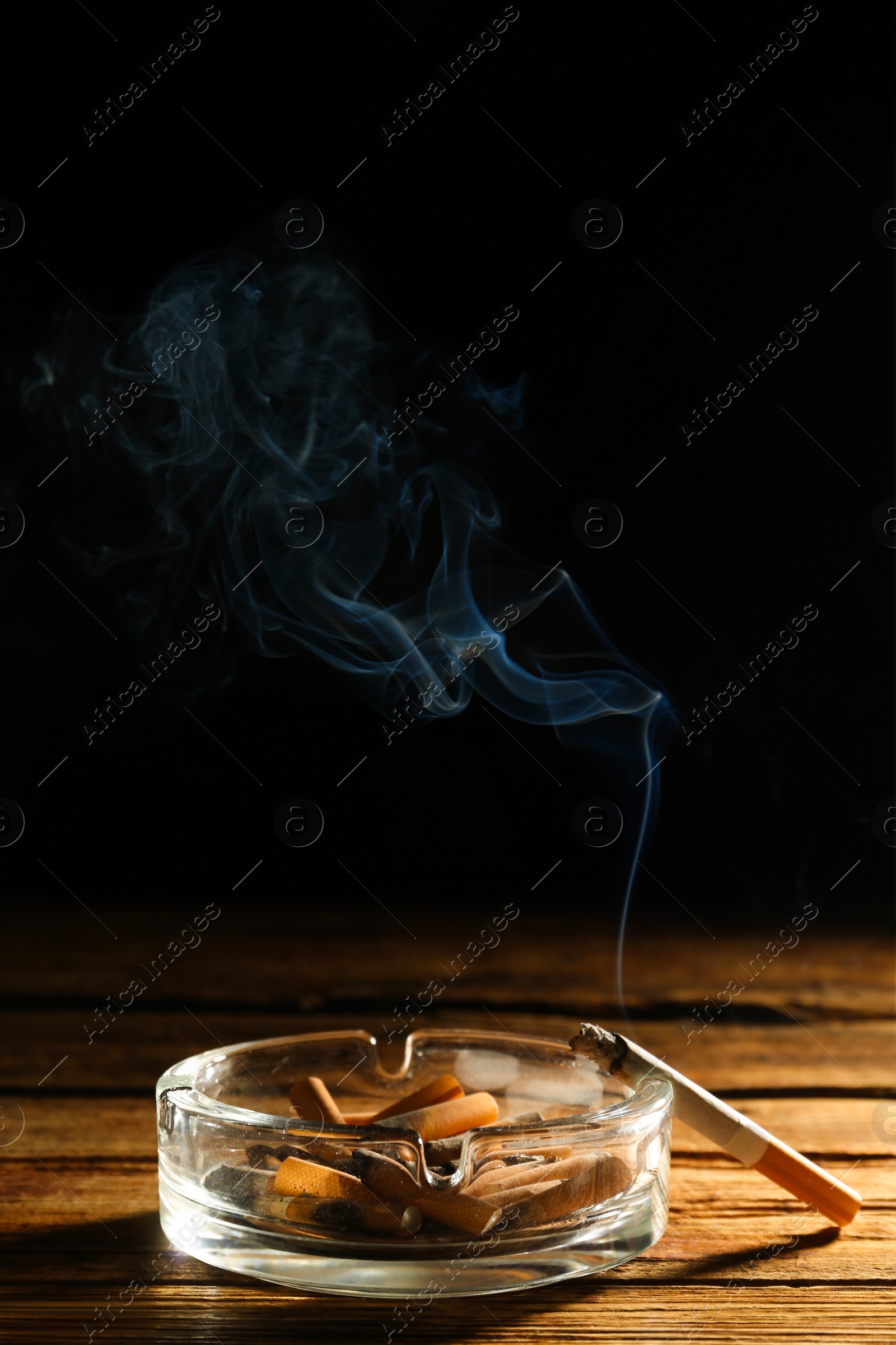 Photo of Glass ashtray with stubs and smoldering cigarette on wooden table against black background. Space for text