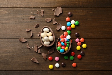 Photo of Broken chocolate egg and colorful candies on wooden table, flat lay