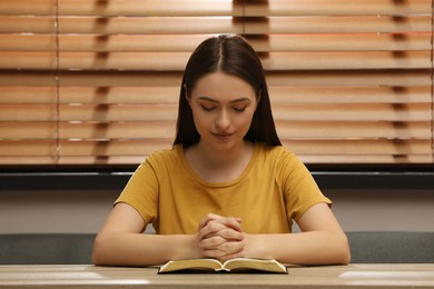 Photo of Religious young woman praying over Bible at wooden table indoors