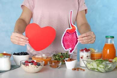 Photo of Woman holding paper intestine and heart near table with food, closeup. Balanced nutrition for healthy digestive system