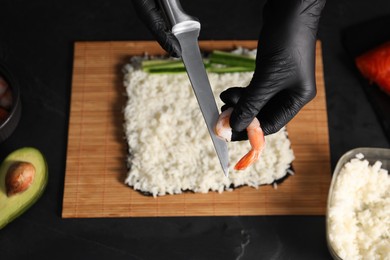 Photo of Chef in gloves with shrimp and knife over sushi roll at dark table, closeup