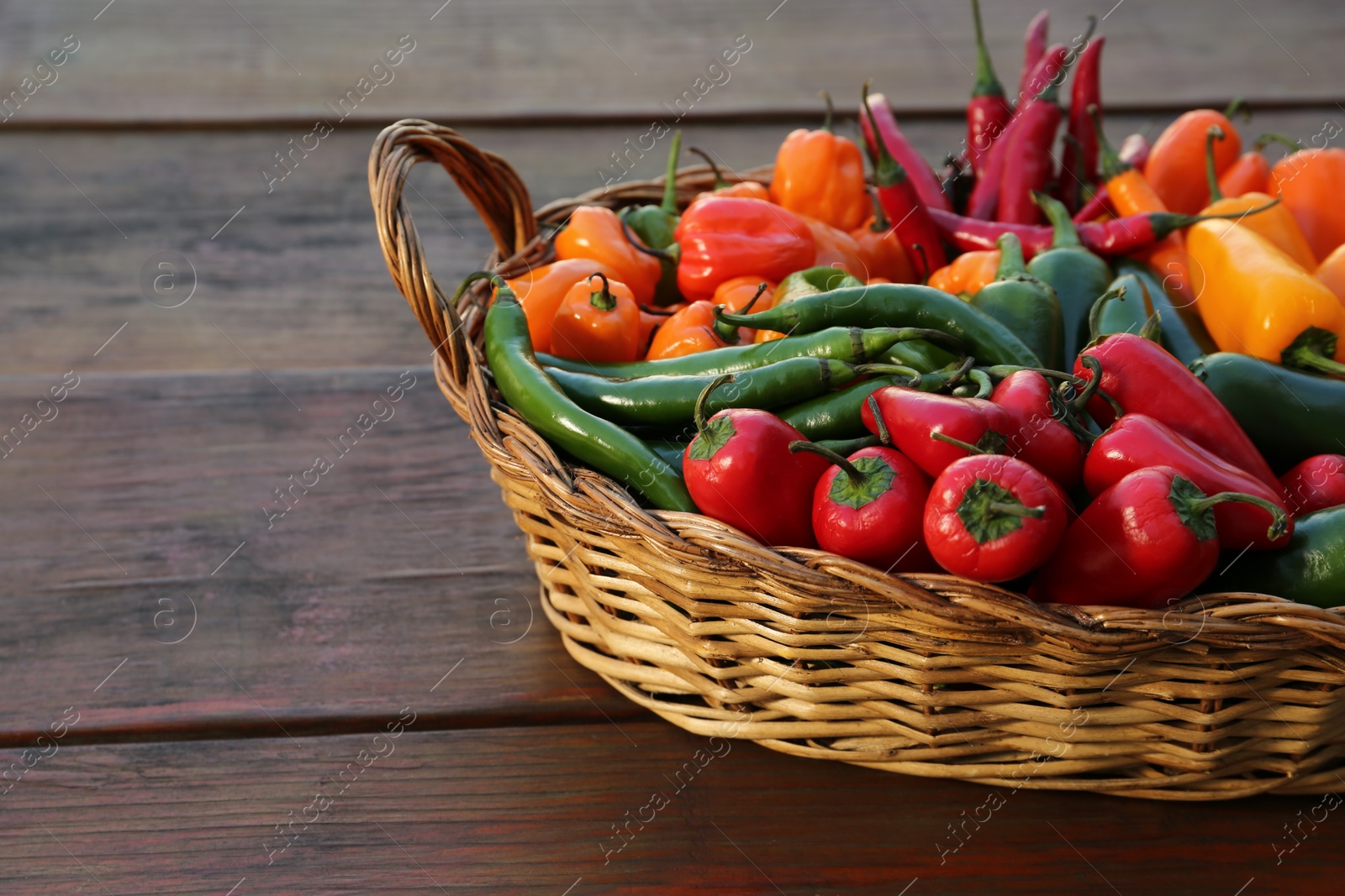 Photo of Wicker basket with many different fresh chilli peppers on wooden table, space for text