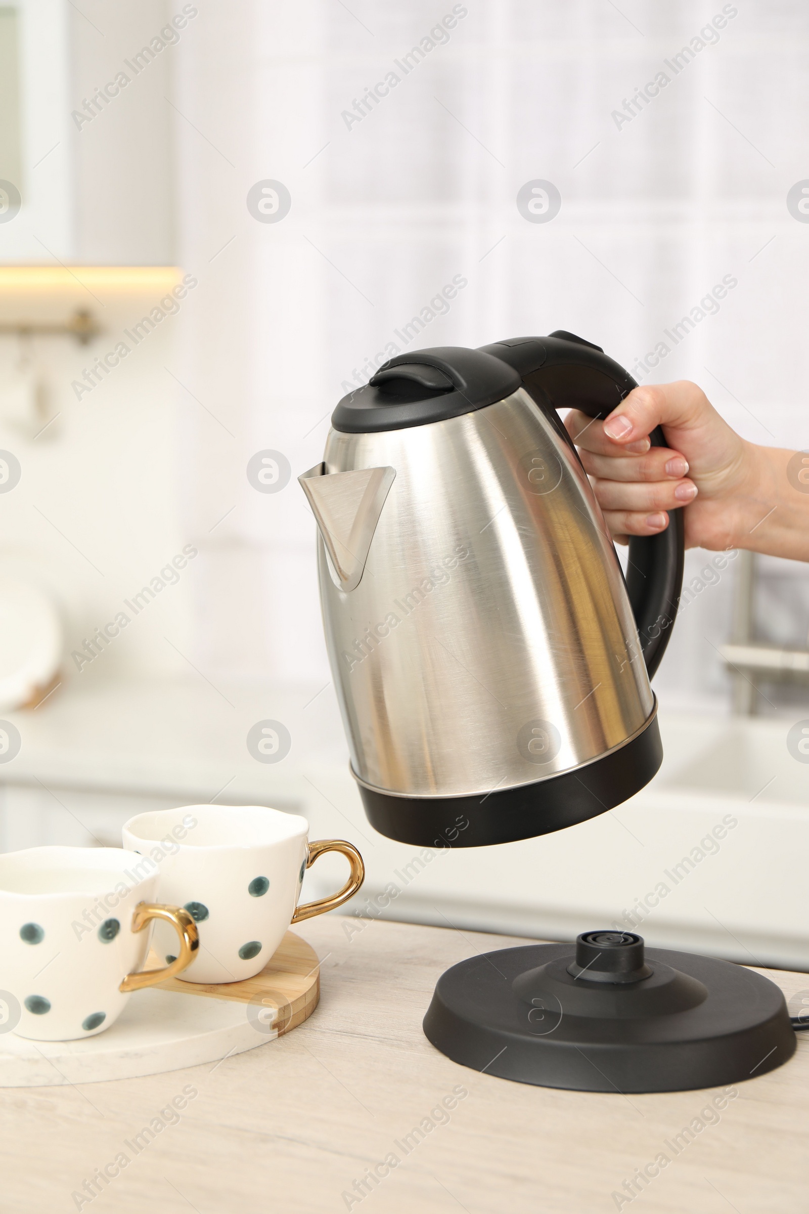 Photo of Woman pouring hot water from electric kettle into cup in kitchen, closeup