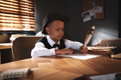 Photo of Cute little detective writing with feather at table in office