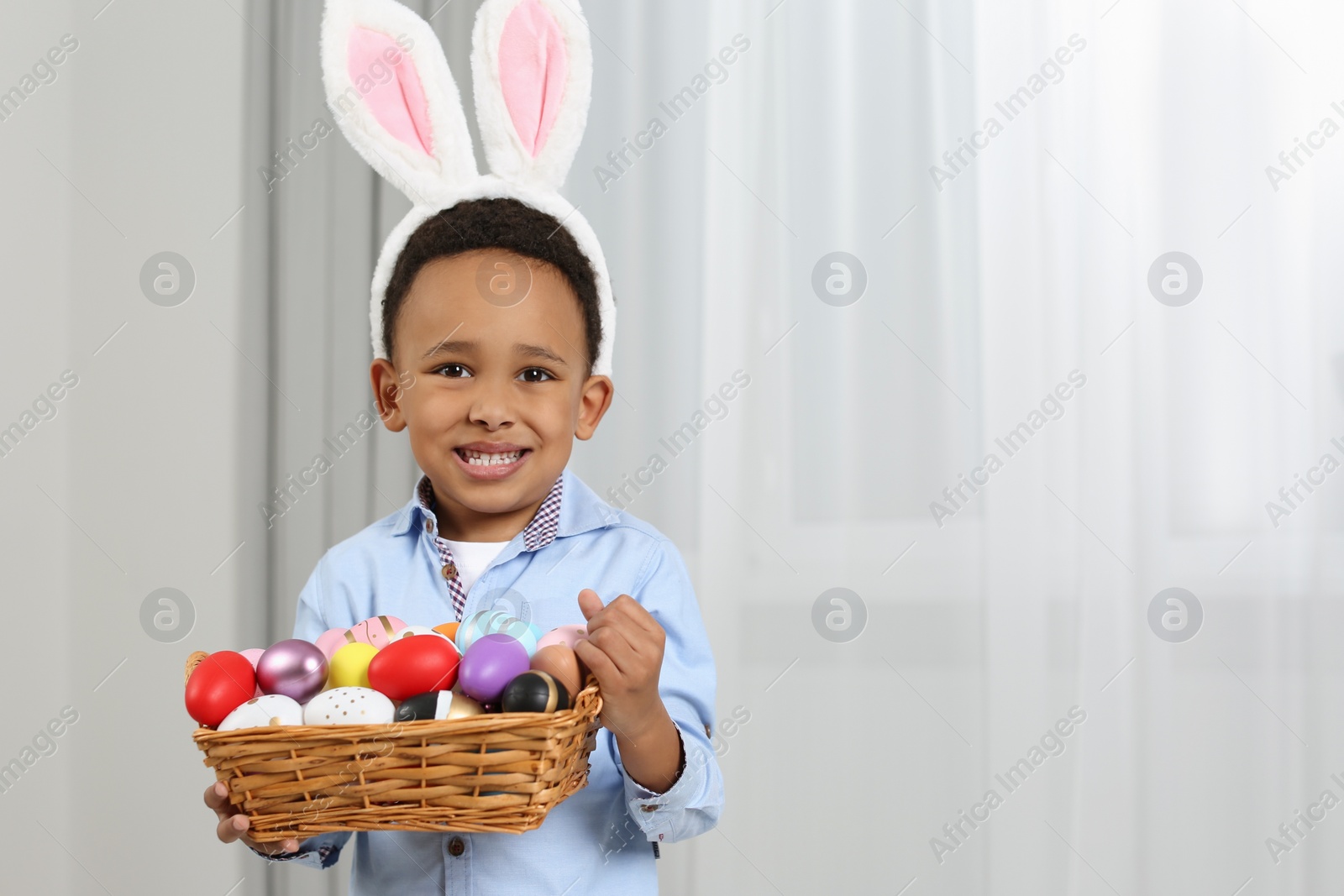 Photo of Cute African American boy in bunny ears headband holding wicker tray with Easter eggs indoors, space for text