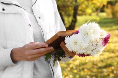 Woman reading book and holding beautiful flowers in park on autumn day, closeup