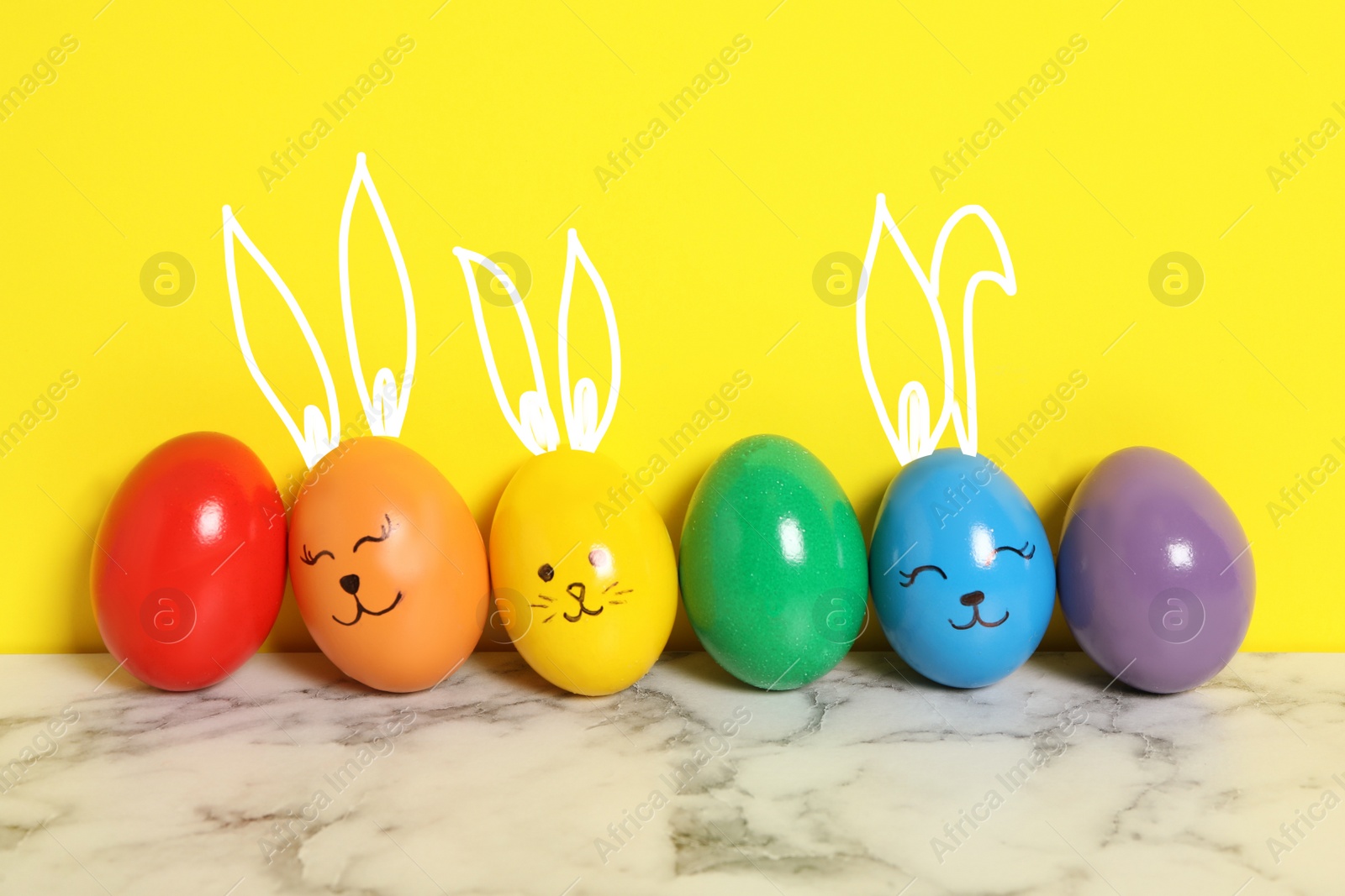 Image of Several eggs with drawn faces and ears as Easter bunnies among others on white marble table against yellow background