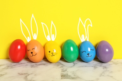 Image of Several eggs with drawn faces and ears as Easter bunnies among others on white marble table against yellow background