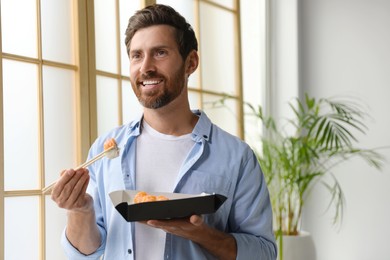 Happy man eating sushi rolls with chopsticks indoors