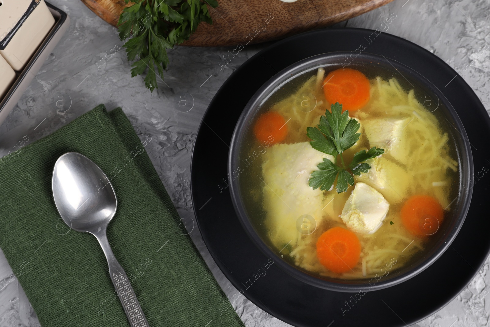 Photo of Tasty chicken soup with noodles, carrot and parsley in bowl served on grey textured table, flat lay