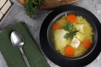 Tasty chicken soup with noodles, carrot and parsley in bowl served on grey textured table, flat lay