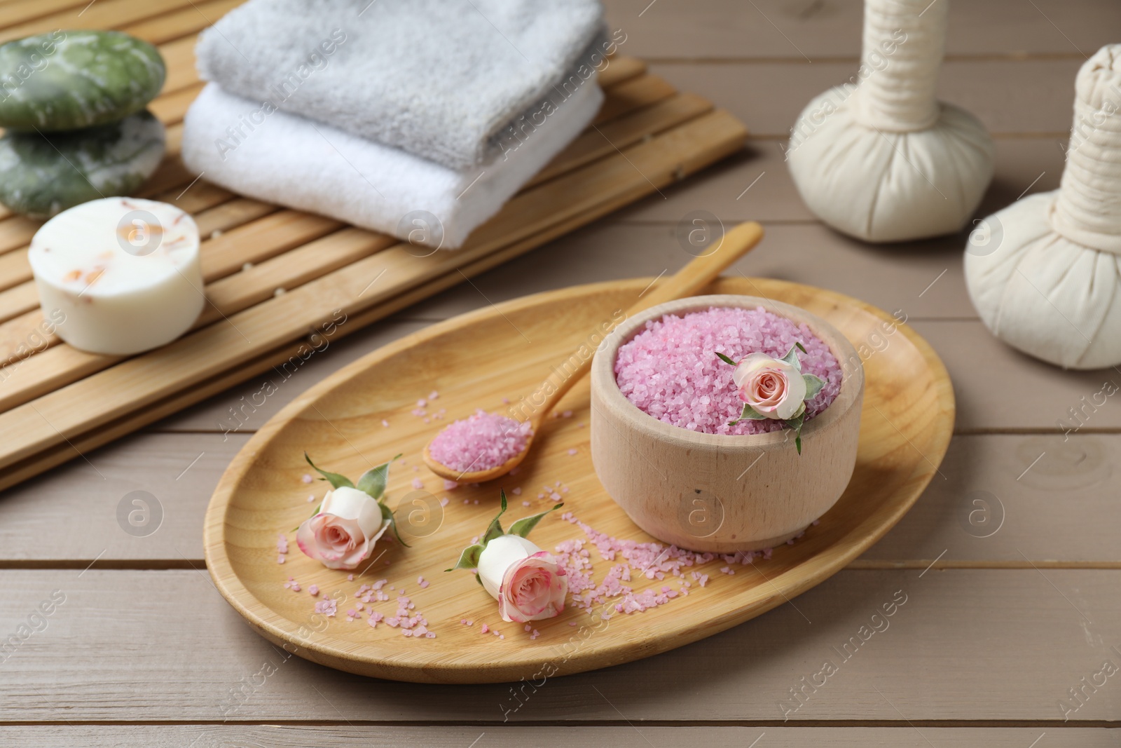 Photo of Bowl of pink sea salt, roses, herbal massage bags and towels on wooden table