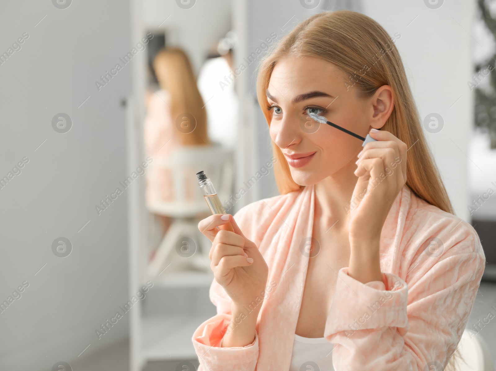 Photo of Young woman applying oil onto her eyelashes near mirror indoors