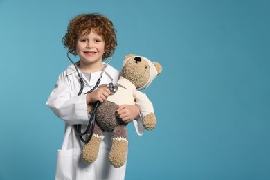Photo of Little boy in medical uniform examining toy bear with stethoscope on light blue background. Space for text