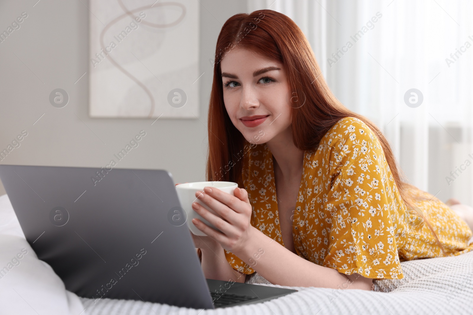 Photo of Beautiful woman with cup of drink and laptop on bed in bedroom