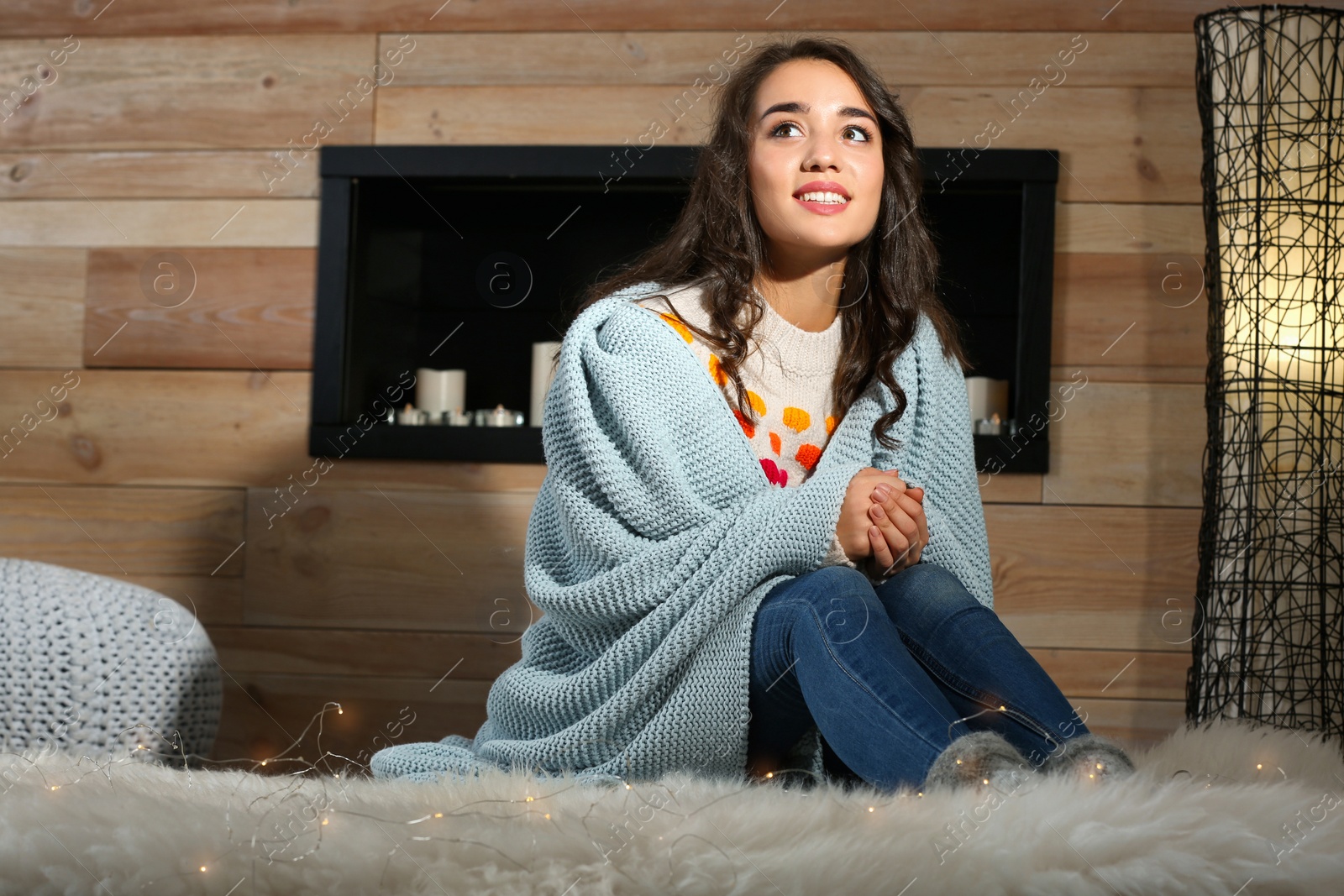 Photo of Young beautiful woman in warm sweater with plaid sitting on fluffy rug at home